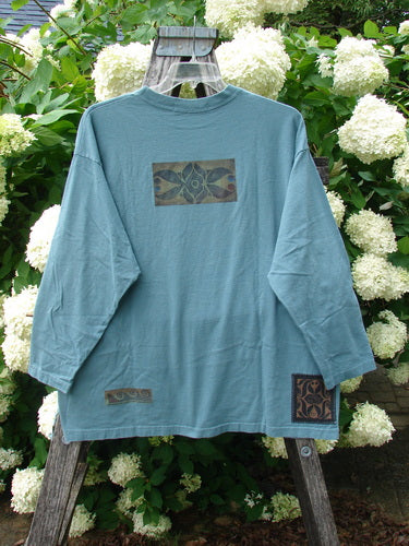 Bluefishfinder.com 1997 Long Sleeved Tee Double Celtic Flower Cypress Size 2 in faded cypress blue, a relaxed long-sleeved cotton shirt with a crew neck and soft, worn texture. It hangs from a wooden ladder before white hydrangeas, with Celtic flower patches on the chest and hem.