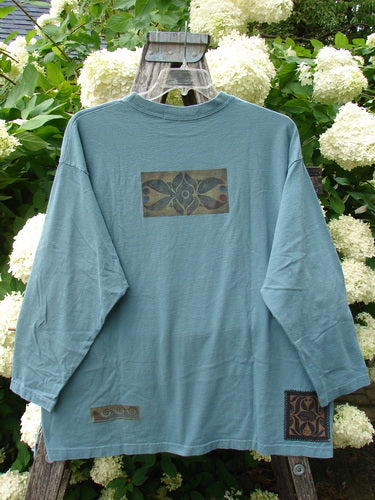 Bluefishfinder.com 1997 Long Sleeved Tee Double Celtic Flower Cypress Size 2 in muted blue-green cotton, with a relaxed crew neck and long sleeves. A rectangular Celtic flower graphic sits at the chest, with smaller matching patches near the hem, hanging against white hydrangeas and weathered wood.