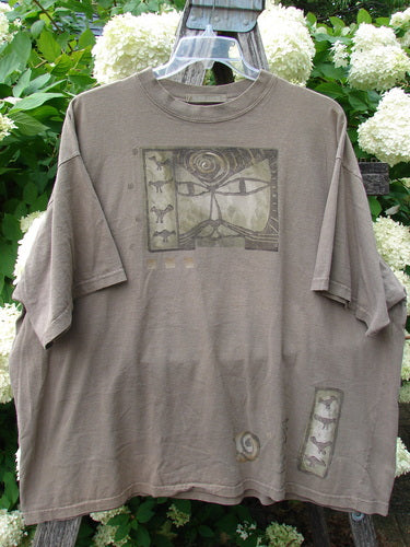 Bluefishfinder.com 1995 Short Sleeved Tee Kitty Kitty Bramble Size 2 in muted brown-gray cotton, with a loose short-sleeve cut, faded graphic panels, and a soft worn finish. The tee hangs on a clear hanger against leafy white hydrangeas, creating a natural, rustic backdrop.