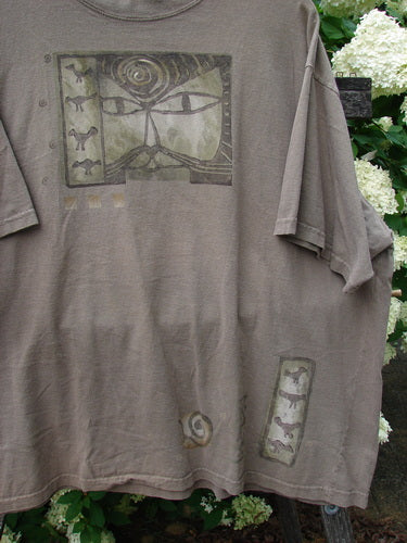 Bluefishfinder.com 1995 Short Sleeved Tee Kitty Kitty Bramble Size 2 in muted taupe, a relaxed short-sleeve shirt with a faded graphic print on the chest and a small vertical motif near the hem. The soft, washed cotton drapes loosely, with the garment shown against leafy white flowers.