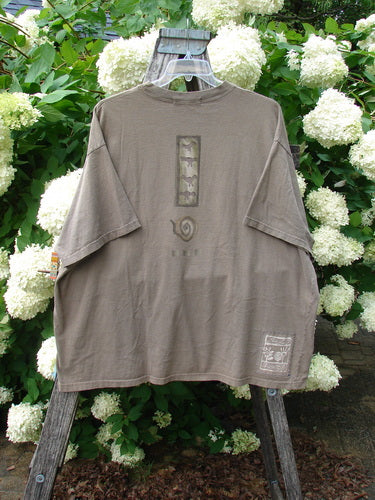 Bluefishfinder.com 1995 Short Sleeved Tee Kitty Kitty Bramble Size 2 in muted taupe, a loose short-sleeve T-shirt with a faded front graphic, stitched hems, and a small printed label near the lower hem. It hangs from a wooden ladder among white hydrangeas, with soft outdoor light and earthy greens framing the worn cotton texture.
