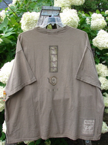 Bluefishfinder.com 1995 Short Sleeved Tee Kitty Kitty Bramble Size 2 in muted taupe, a loose short-sleeved T-shirt with a faded graphic panel at the center chest and a small printed patch near the lower hem. It hangs from a clear hanger against white hydrangeas and green foliage, giving the cotton fabric a soft, worn matte finish.