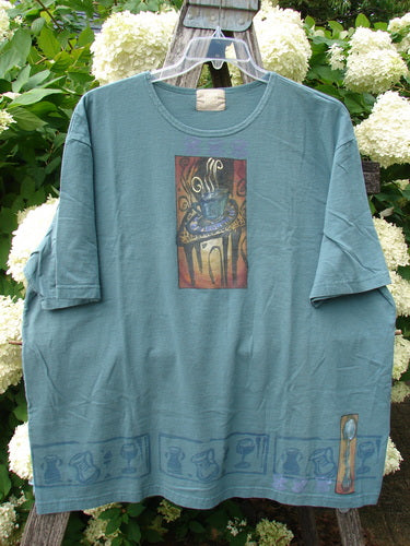 Bluefishfinder.com 2000 Artist Choice Short Sleeved Tee Steaming Teacup Green Mineral Size 2 in soft green mineral cotton, with a relaxed short-sleeve cut and a rectangular steaming teacup graphic centered on the chest. A muted border of repeated teacup motifs runs along the hem, with natural folds and outdoor light revealing the tee’s worn, matte texture.