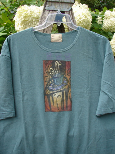 Bluefishfinder.com 2000 Artist Choice Short Sleeved Tee in green mineral, size 2, with a soft crew neck and short sleeves. A centered steaming teacup graphic sits on the faded cotton front, photographed on a clear hanger against leafy white hydrangeas.