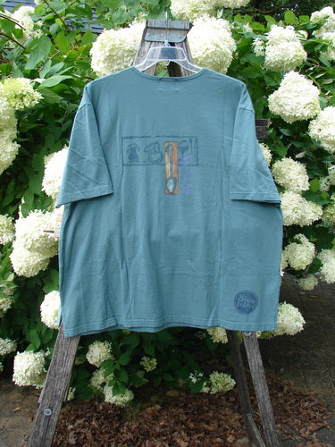 Bluefishfinder.com 2000 Artist Choice Short Sleeved Tee in green mineral, size 2, shown as a soft short-sleeve T-shirt with a faded graphic of a steaming teacup at the chest and a small circular print near the hem. The relaxed tee hangs from a wooden ladder amid white hydrangea blooms, with muted blue-green fabric and weathered, natural textures.