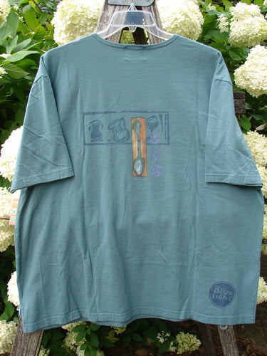 Bluefishfinder.com 2000 Artist Choice Short Sleeved Tee in steaming teacup green, a relaxed cotton T-shirt with a faded graphic centered on the chest and a small circular logo near the hem. It hangs on a clear hanger against leafy white flowers.