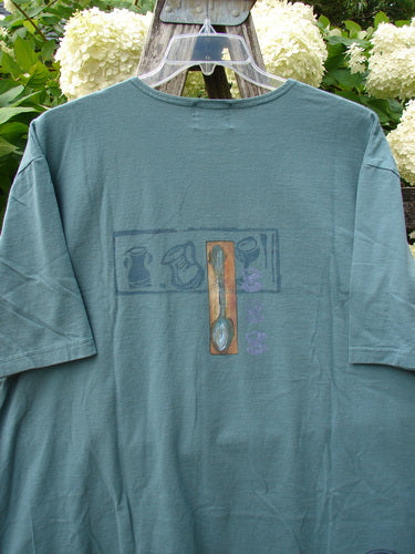 Bluefishfinder.com 2000 Artist Choice Short Sleeved Tee Steaming Teacup Green Mineral Size 2, a green mineral short-sleeved T-shirt with a faded central steaming teacup graphic. The soft cotton jersey hangs from a hanger, with the muted print and washed texture set against leafy greenery and white blooms.