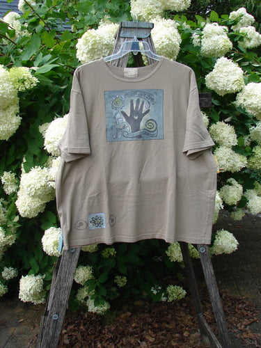 Bluefishfinder.com 2000 Artist Choice Short Sleeved Tee Earth Day Deep Dove Size 2 in muted gray-green cotton, with a square chest graphic of a raised hand and spiral motifs and a small Earth Day-style emblem near the hem. The tee hangs on a wooden ladder against dense white hydrangeas and green foliage.