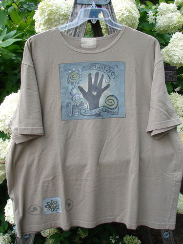 Bluefishfinder.com 2000 Artist Choice Short Sleeved Tee Earth Day Deep Dove Size 2 in muted taupe cotton, with a loose short-sleeve cut and a faded square Earth Day graphic centered on the chest. A small matching motif near the hem adds detail, while the tee hangs on a clear hanger against dense green foliage and white flowers.