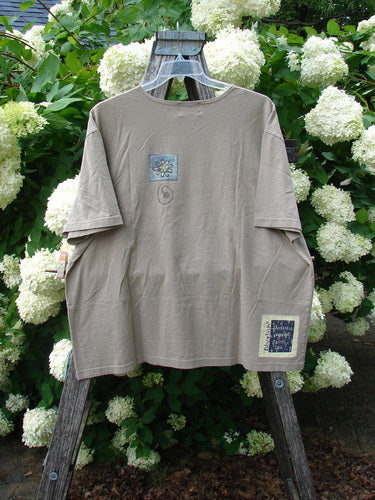 Bluefishfinder.com 2000 Artist Choice Short Sleeved Tee Earth Day Deep Dove, size 2, in muted gray-brown cotton with a loose crewneck silhouette and short sleeves. Small graphic patches sit on the chest and lower hem, while the tee hangs from a wooden easel amid white hydrangeas.