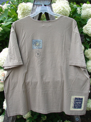 Bluefishfinder.com 2000 Artist Choice Short Sleeved Tee Earth Day Deep Dove Size 2 in muted taupe, a short-sleeved cotton T-shirt with a relaxed drape, small chest graphic, and square lower-right patch. It hangs on a clear hanger against white hydrangeas and green leaves.
