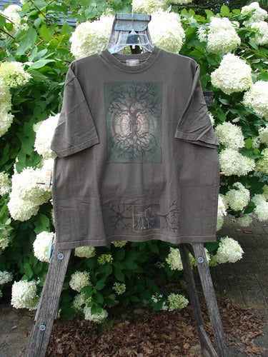 Bluefishfinder.com 2000 Short Sleeved Tee Earthen Tree Domino Size 2 in muted brown-gray cotton, with a relaxed short-sleeve cut and faded tree graphic on the chest. It hangs on a wooden stand before white blossoms and dense green foliage, creating a natural, earthy contrast.