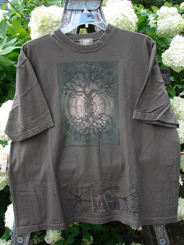 Bluefishfinder.com 2000 Short Sleeved Tee Earthen Tree Domino Size 2 in faded charcoal cotton, with a loose short-sleeve cut and a centered tree graphic in muted green and brown. It hangs front-facing against leafy white blossoms, its soft, worn texture and matte finish visible in the natural light.