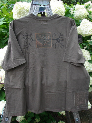 Bluefishfinder.com 2000 Short Sleeved Tee Earthen Tree Domino Size 2 in muted gray-brown cotton, shown front-facing with short sleeves, a faded tree graphic across the chest, and a small square print near the hem. The tee hangs on a clear hanger against white flowers and green foliage.