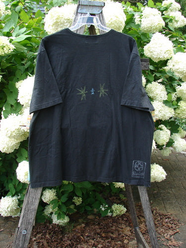 Bluefishfinder.com 1999 Short Sleeved Tee Geisha Gal Black Size 2, a black short-sleeved T-shirt with a loose, boxy silhouette and faint green graphic marks centered on the chest. It hangs from a wooden ladder in front of white hydrangeas and dense green foliage, with a small label near the lower hem.