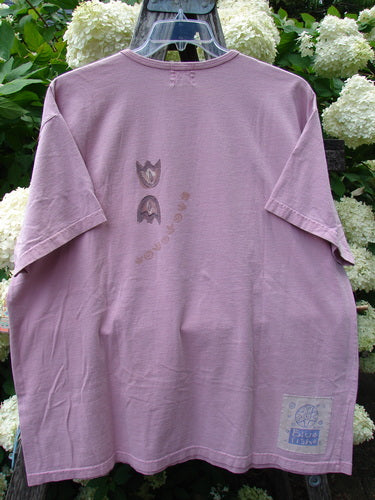 Bluefishfinder.com 1999 Short Sleeved Tee Double Daisy Peony Size 2 in dusty pink, a short-sleeved T-shirt with a relaxed drape, small chest graphic, and pale patch at the lower hem. The soft fabric hangs from a hanger against white hydrangeas, with muted green foliage framing the scene.
