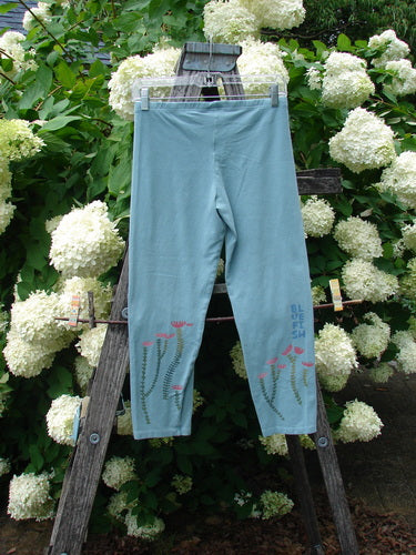 Bluefishfinder.com Barclay Cotton Lycra Crop Legging Rising Floral Sky Size 0 in pale sky blue, shown as a cropped legging with a smooth stretch finish and floral vine graphics near the lower legs. It hangs centered on a wooden ladder amid white hydrangea blooms, with soft natural light and a fresh garden palette.