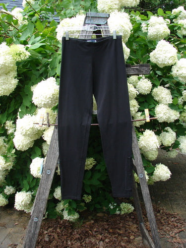 Bluefishfinder.com Barclay Cotton Lycra Side Gather Legging in unpainted black, size 0, shown as a sleek black pair with a smooth stretch finish and side-gather detailing. It hangs centered on a wooden ladder, framed by dense white hydrangea blooms and green foliage in soft outdoor light.