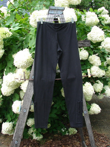 Bluefishfinder.com Barclay Cotton Lycra Side Gather Legging in unpainted black, size 0, shown as a slim, ankle-length pair with a smooth stretch finish and subtle side-gather detail. The leggings hang centered on a wooden ladder, framed by dense white hydrangea blooms and green foliage.