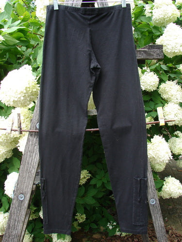 Bluefishfinder.com Barclay Cotton Lycra Side Gather Legging in unpainted black, size 0, shown as a pair of slim, full-length leggings with a smooth matte finish and gathered side seams. The dark fabric hangs against a rustic wooden support, framed by dense white hydrangea blooms and green foliage.