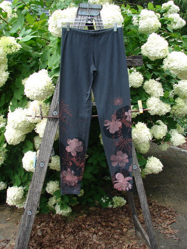 Bluefishfinder.com Barclay Cotton Lycra Legging Daisy Twig in off black, size 0, with a slim cropped silhouette and pink daisy-and-twig print near the lower legs. The matte stretch fabric hangs from a wooden ladder outdoors, against white hydrangeas and dense green foliage.