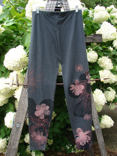 Bluefishfinder.com Barclay Cotton Lycra Legging Daisy Twig Off Black, size 0, in a dark charcoal stretch knit with pink floral twig prints concentrated at the lower legs. The slim leggings hang centered on a wooden ladder, framed by white hydrangeas and green foliage in soft outdoor light.