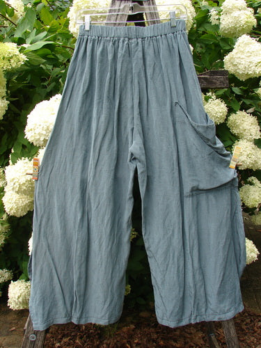 Bluefishfinder.com Barclay Silk Dimension Pant Unpainted Dark Sage Size 1, wide-legged silk pants in muted dark sage with a softly wrinkled texture and gathered elastic waist. A draped side pocket panel falls along one leg, while the garment hangs centered against dense white hydrangeas and green foliage.
