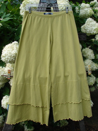 Bluefishfinder.com Barclay Cotton Lycra Crop 2 Petal Pantaloon in unpainted sunshine, size 0, shown as olive-green cropped pants with a soft stretch knit, elastic waistband, and layered petal-like hems. The fabric hangs in a straight, fluid drape against a backdrop of white flowers and dark greenery.