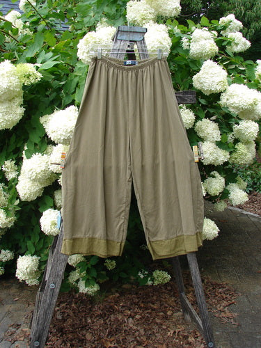 Bluefishfinder.com Barclay lyocell flutter bottom wide leg pant in unpainted yellow olive, size 0, shown as loose olive-toned trousers with a gathered elastic waist and wide, draping legs. The lightweight lyocell fabric has a soft matte finish and fluttered hems, hanging from a wooden ladder against white hydrangeas and green foliage.