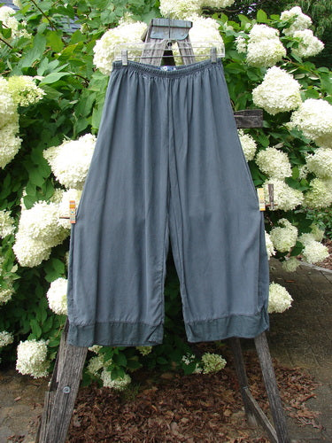 Bluefishfinder.com Barclay Lyocell Flutter Bottom Wide Leg Pant in Unpainted Light Storm, size 0, hangs from a rustic wooden ladder in a soft blue-gray tone. The wide legs fall in fluid lyocell folds with fluttered hems, framed by dense white hydrangeas and green foliage.