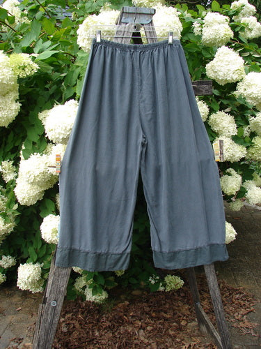 Bluefishfinder.com Barclay Lyocell Flutter Bottom Wide Leg Pant in Unpainted Light Storm, Size 0, shown in a slate-gray drape with a wide, flowing leg and fluttered hem. The lyocell fabric hangs softly from a gathered waistband, framed against white hydrangeas and weathered wood.
