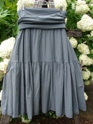 Bluefishfinder.com Barclay Linen Fold Over Single Tier Trinity Skirt in unpainted diatom, size 0, with a soft linen texture, folded waistband, and single tier of gathered fabric that falls into a full, gently flared silhouette. The muted gray-blue skirt hangs on a hanger against leafy white hydrangeas.