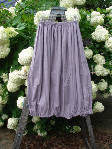 Bluefishfinder.com Barclay Batiste Airy Lawn Skirt in unpainted lavender, size 0, hangs from a wooden ladder in soft outdoor light. The lightweight skirt has a gathered elastic waist, flowing A-line drape, and crinkled lawn texture, set against dense green foliage and white hydrangea blooms.