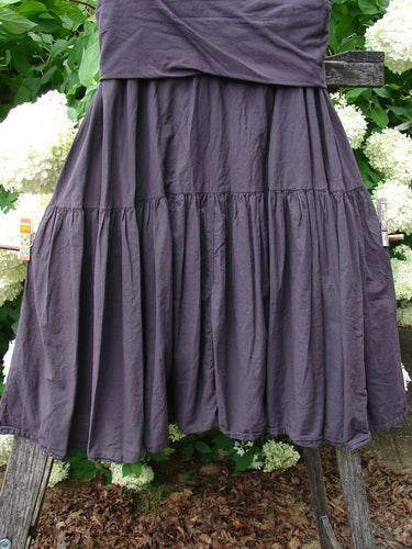 Bluefishfinder.com Barclay Linen Fold Over Single Tier Trinity Skirt in unpainted plum wine, size 0, with a wide fold-over waistband and a single gathered tier that falls in soft, crinkled linen-like drape. The deep purple skirt fills the frame against leafy greenery and white blooms.