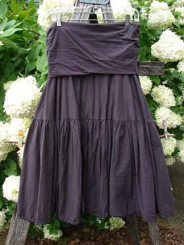 Bluefishfinder.com Barclay Linen Fold Over Single Tier Trinity Skirt in unpainted plum wine, size 0, with a wide fold-over waistband and a single gathered tier that falls into a softly flared linen silhouette. The dark plum fabric hangs against leafy white hydrangeas and wood, emphasizing its textured drape and matte finish.