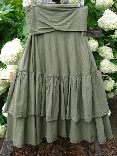 Bluefishfinder.com Barclay Linen Fold Over Two Tier Trinity Skirt in unpainted dried moss, a layered linen skirt with a wide folded waistband and two tiered, softly gathered panels. The muted olive fabric hangs in full, textured folds against a leafy outdoor backdrop.