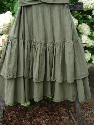 Bluefishfinder.com Barclay Linen Fold Over Two Tier Trinity Skirt in unpainted dried moss, size 0, with a soft linen texture and a layered two-tier silhouette. The full skirt hangs in gathered folds, the muted olive tone set against leafy greenery and white flowers.