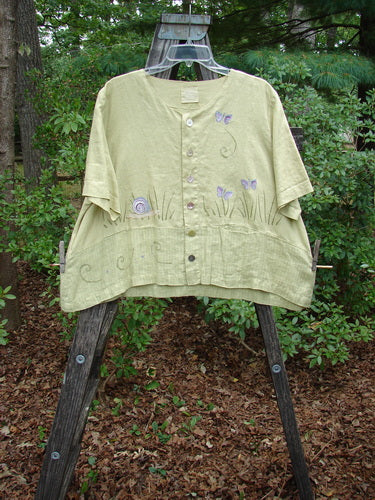 1999 Cabana Top Butterfly Grass Citron Size 2: A lightweight, textured linen shirt with floral and butterfly designs, shell buttons, varying hemline, and a sweet drop pocket, displayed on a hanger outdoors.
