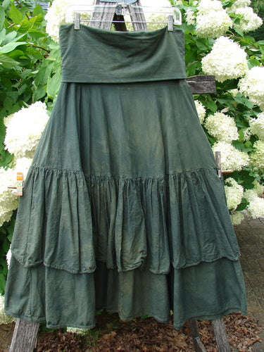 Bluefishfinder.com Barclay Linen Fold Over Two Tier Trinity Skirt in mottled army green, a layered linen skirt with a wide fold-over waistband and two gathered tiers. The softly crinkled fabric hangs in dark, muted folds against a backdrop of white hydrangeas and green foliage.