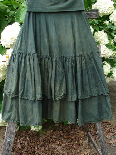 Bluefishfinder.com Barclay Linen Fold Over Two Tier Trinity Skirt in unpainted mottled army, a layered linen skirt with a folded waistband and two gathered tiers. The dark green fabric hangs in soft, textured folds, filling the frame against white flowers and weathered wood.