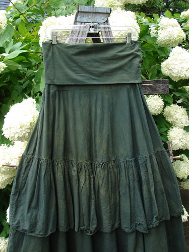 Bluefishfinder.com Barclay Linen Fold Over Two Tier Trinity Skirt, unpainted mottled army green size 0, hangs from a wooden hanger with a folded-over waistband and two tiered, softly gathered linen panels. The dark, textured fabric is shown against leafy white hydrangeas.