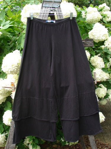 Bluefishfinder.com Barclay Cotton Lycra Crop 2 Petal Pantaloon in unpainted black, size 0, hangs from a clear hanger in a full-length view. The soft cotton-lycra fabric falls in a loose, tiered silhouette with a wide hem and matte finish, framed by white hydrangeas and weathered wood.