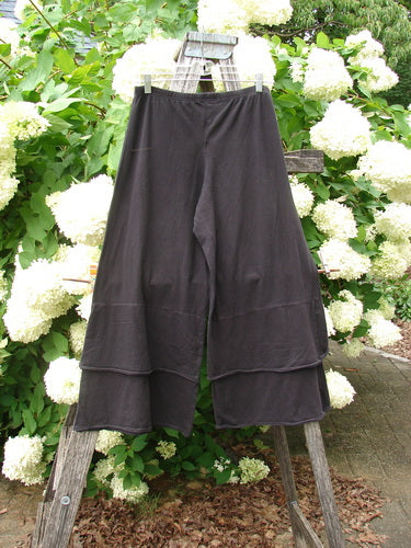 Bluefishfinder.com Barclay Cotton Lycra Crop 2 Petal Pantaloon in unpainted black, size 0, shown as wide, layered cropped pants with a soft stretch knit and flowing petal-shaped hems. The dark fabric hangs from a rustic wooden ladder amid dense white hydrangeas and green leaves.
