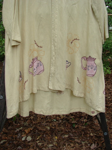 1999 Cream and Sugar Coat Pitcher Plantain OSFA on a rack, featuring teapot and pitcher drawings, medium-weight linen, oversized shell button, deep side pockets, and a unique upward scoop front hem.