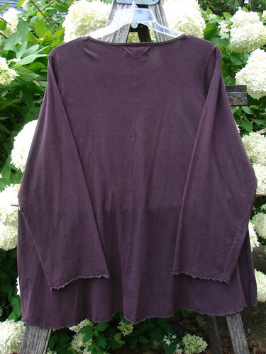 Bluefishfinder.com Barclay Long Sleeved Studio Cafe Tee in Celestial Purple Plum, size 2, a long-sleeved tee with a wide crew neckline, relaxed drape, and raw, slightly frayed hems. The deep plum knit hangs against leafy white hydrangeas, creating a soft, layered contrast.