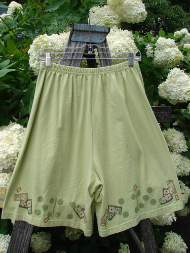 Bluefishfinder.com 1998 Boxer Short Continuous Domino Clover Size 1 in pale olive cotton, hanging from a wooden ladder with clear clips. The loose boxy shorts have a soft matte finish and a hem printed with small domino and clover motifs, set against white hydrangeas and dark green foliage.