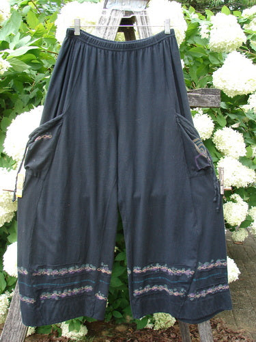 Bluefishfinder.com Barclay Crop Tie Pocket Pinch Pant Wind Chain in black, size 0, hangs from a clip hanger with a relaxed cropped silhouette and gathered waistband. The dark fabric is accented by tie pockets and delicate chain-like trim near the hems, set against bright white flowers and green foliage.