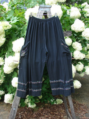 Bluefishfinder.com Barclay Crop Tie Pocket Pinch Pant Wind Chain in black, size 0, with a relaxed cropped silhouette, gathered tie waist, and side pockets. The dark fabric is trimmed with subtle patterned bands near the hems, photographed against lush white hydrangeas and green foliage.