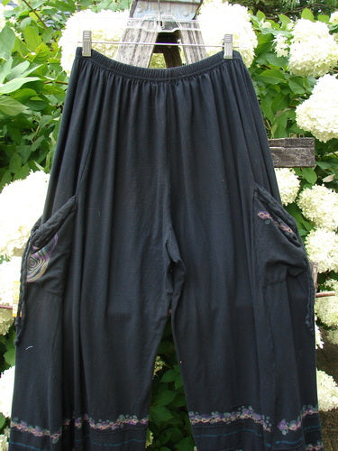 Bluefishfinder.com Barclay Crop Tie Pocket Pinch Pant Wind Chain in black, size 0, with a gathered elastic waist and wide cropped legs. The soft drape hangs from clips, while side tie pockets and subtle patterned trim near the hems add texture against the leafy backdrop.