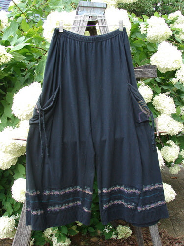 Bluefishfinder.com Barclay Crop Tie Pocket Pinch Pant Wind Chain Black Size 0, a pair of black cropped pants with a gathered waist, side tie pockets, and soft drape. Decorative floral bands edge the hem, while the garment hangs centered against leafy white blossoms and weathered wood.